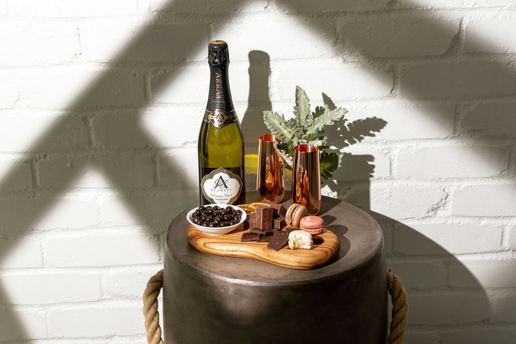 A bottle of Australian sparkling wine paired with copper flutes accompanied by a small dessert platter of chocolate, fudge, nougat and other sweet treats photographed with some window shadow lines for effect.
