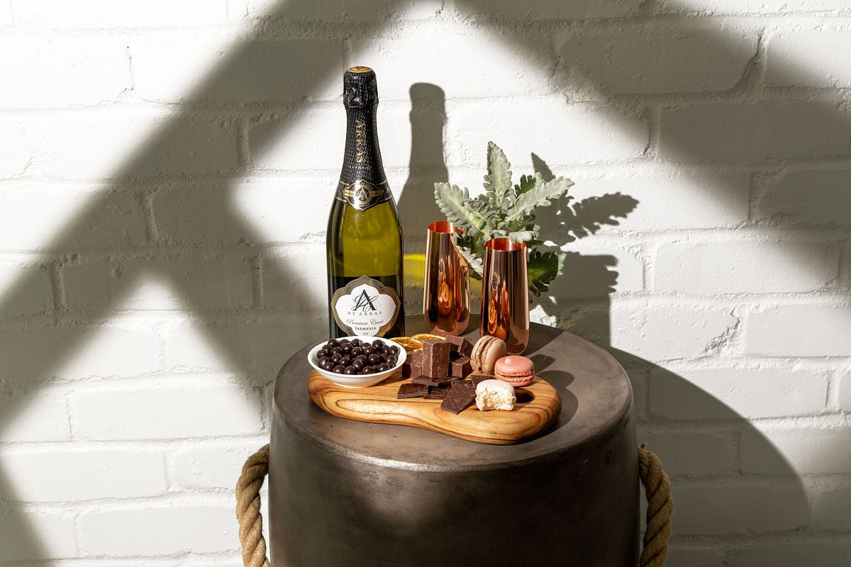 celebration gift hamper - bottle of champagne, copper champagne flutes, premium chocolate on display on timber serving board