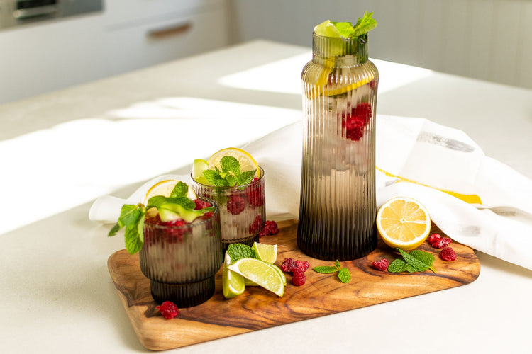 Mouthblown, ribbed glass decanter and matching glassware pair filled with citrus, berries and mint on a white kitchen bench.