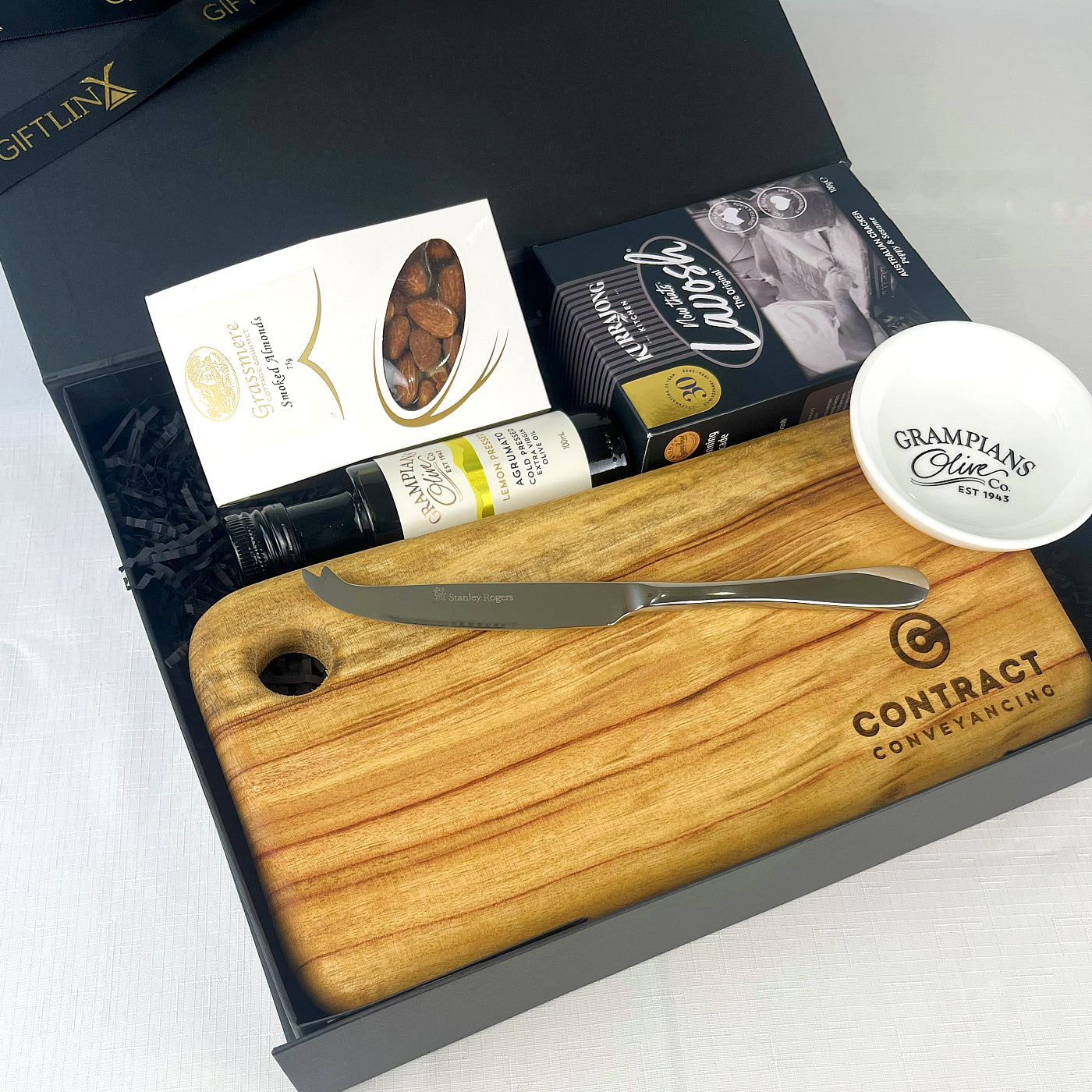 Wooden cutting board with knife, olive oil bottle, and snack in a gift box.