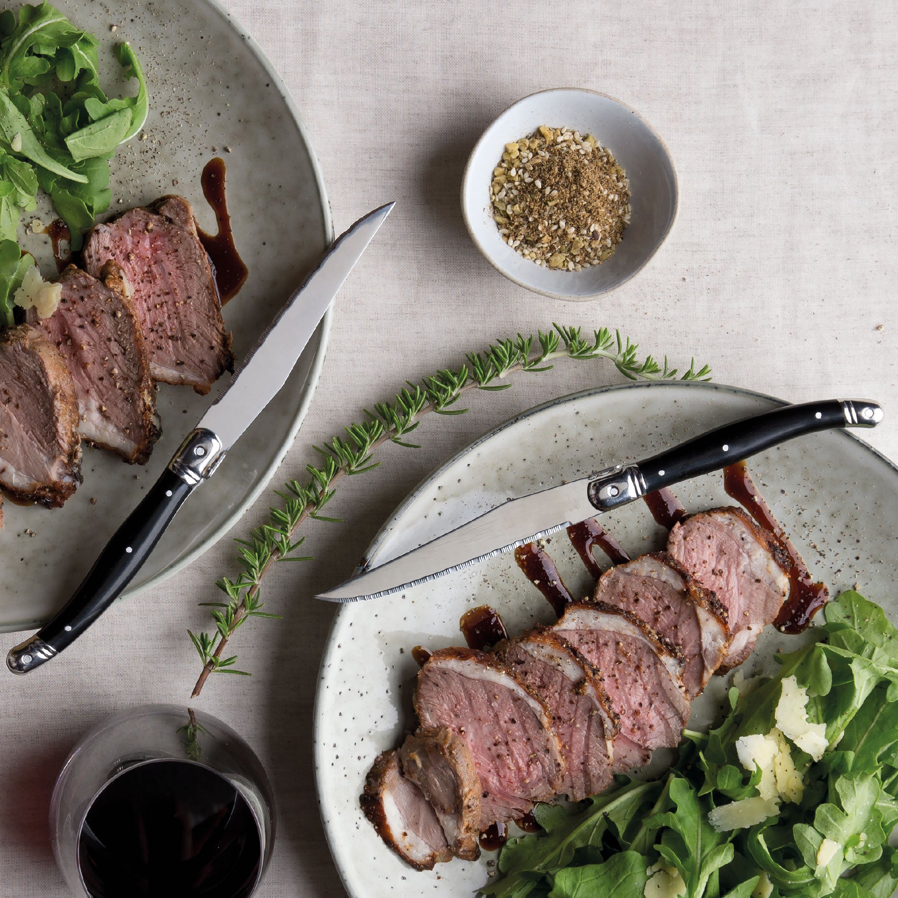 lifestyle photograph of black handled steak knives on plates with sliced meat, herbs and gravy