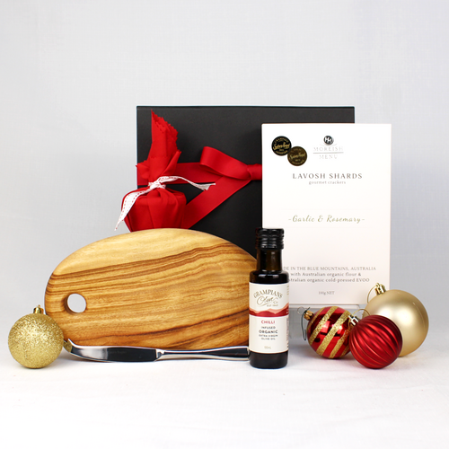 black gift box with red ribbon behind a small timber cheeseboard with single stainless steel knife, infused olive oil, box of lavosh shards and a small Christmas pudding in red fabric surrounded by Christmas baubles