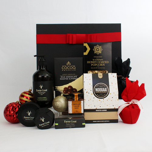 black gift box with red ribbon standing behind a range of Farm Goat bdy wash, shampoo bar and soap alongside 2 christmas puddings in red fabric with jar of pudding sauce, a box of choocolate coated almonds, honey coated popcorn and nougat pieces