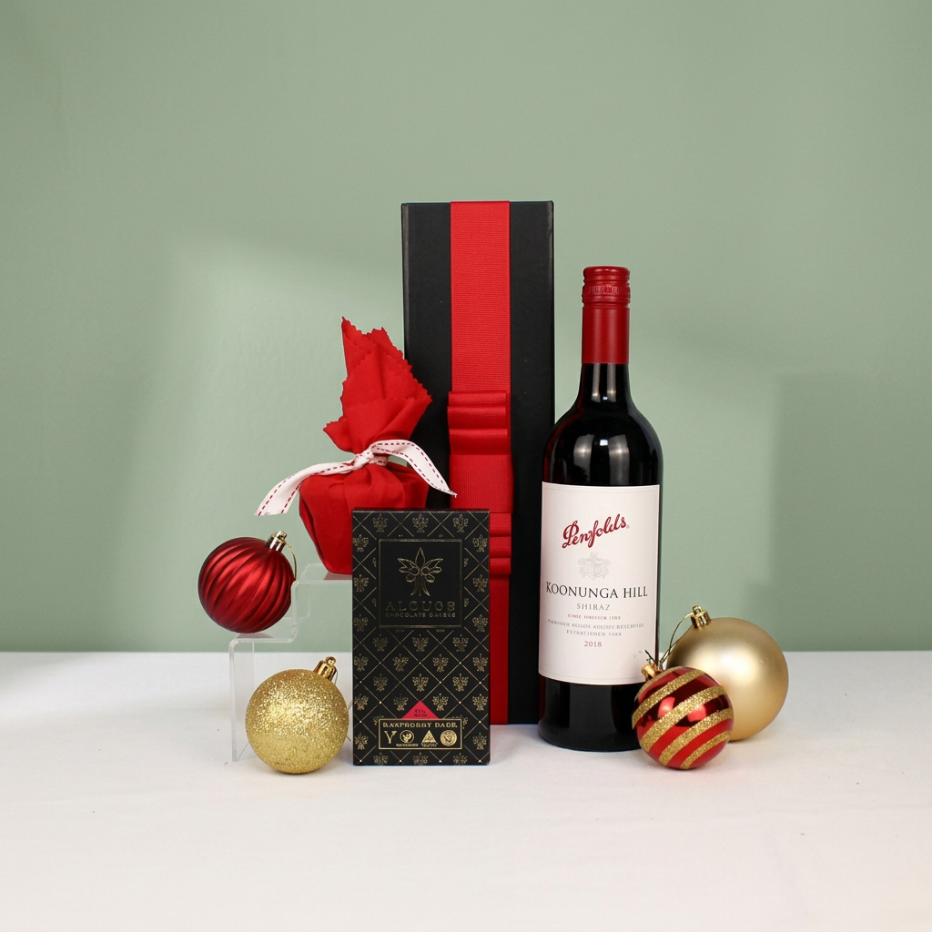 Black gift box with read ribbon behind a bottle of Australian red wine, Christmas pudding and handmade chocolate surrounded by Christmas baubles