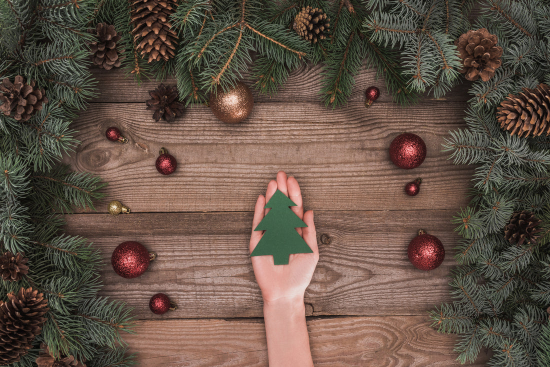 Hand holding a small green christmas tree surrounded by a green garland, red baubles and pine cones