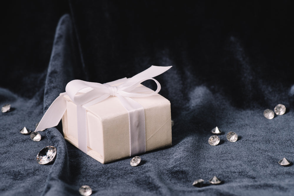 Dark cloth laying on table with a white gift box with white ribbon sitting on top surrounded by scattered diamonds