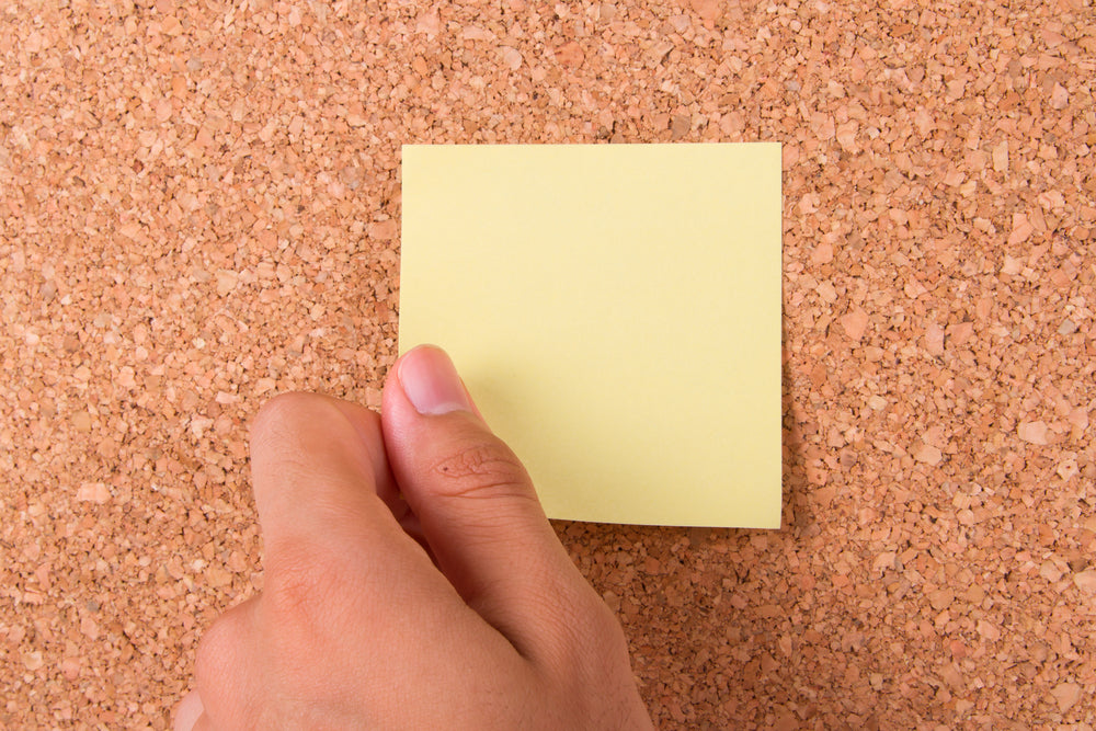 Cork board with a hand placing a yellow post it note on it