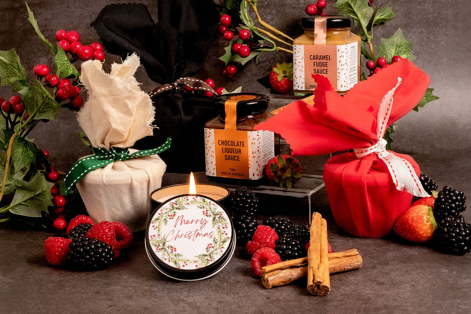 Festive scene with Christmas puddings wrapped in red and natural fabric alongside 2 jars of pudding sauce with a small Christmas candle lit in the foreground