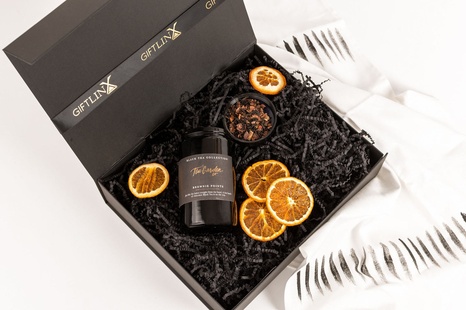 Luxury Black Gift Box with black shred paper and black and gold branded ribbon tied in a bow. Box contains loose leaf tea jar and orange slices.