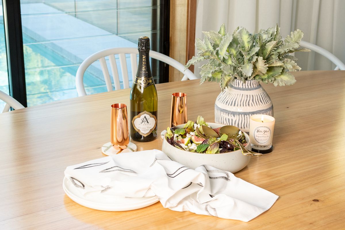 A dining table set with contents from a settlement gift box - with a bottle of sparkling wine, copper champagne flutes on marble coasters, stone salad bowl filled with summer salad and a luxury hand poured soy candle burning on the table.