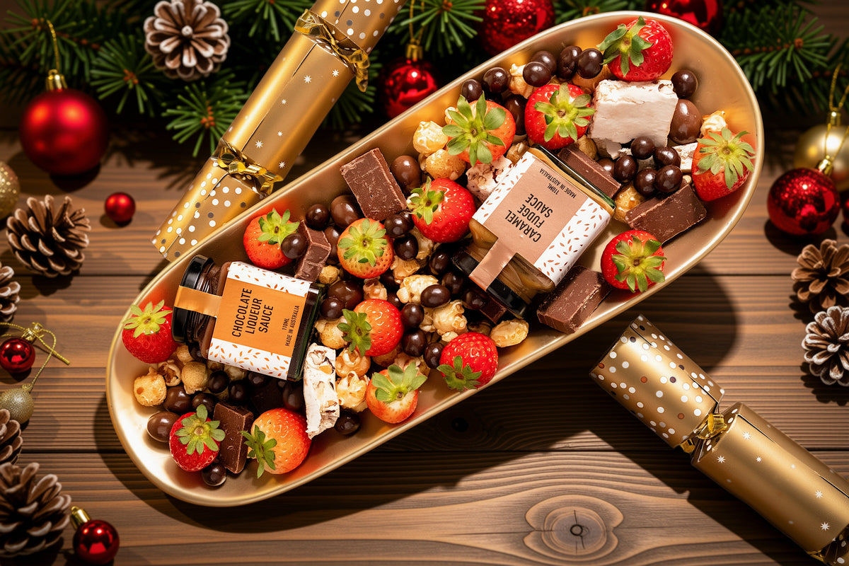 A festive holiday gift tray filled with assorted chocolates, strawberries, popcorn, and small wrapped treats, styled on a wooden surface with Christmas ornaments, pinecones, and gold cracker decorations.