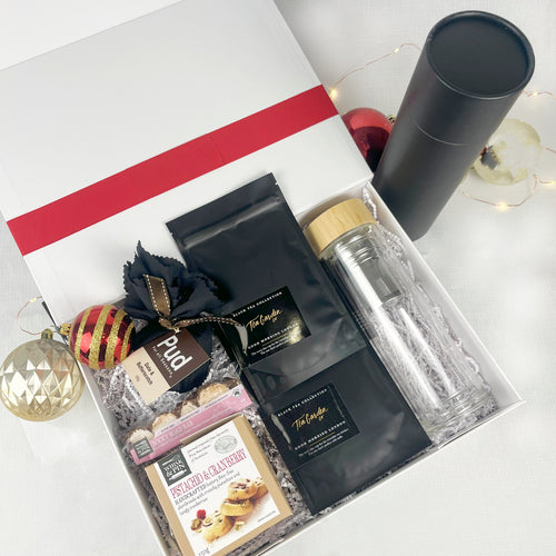 Gift set with a black cylindrical container, glass bottle, tea, shortbread and chocolate on a white surface.