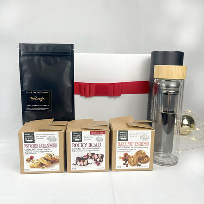 Assorted food products including a black tea sachet, a clear glass flask with a bamboo lid, and three boxes of chocolate  and cookies on a white background.