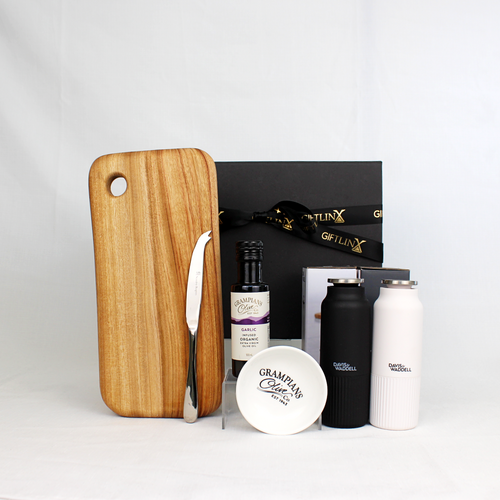 A black and white pair of salt and pepper mills alongside a bottle of infused olive oil with dipping bowl and a handmade cheese board with cheese knife in front of a black gift box with gold ribbon