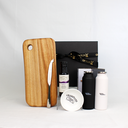 A black and white pair of salt and pepper mills alongside a bottle of infused olive oil with dipping bowl and a handmade cheese board with cheese knife in front of a black gift box with gold ribbon