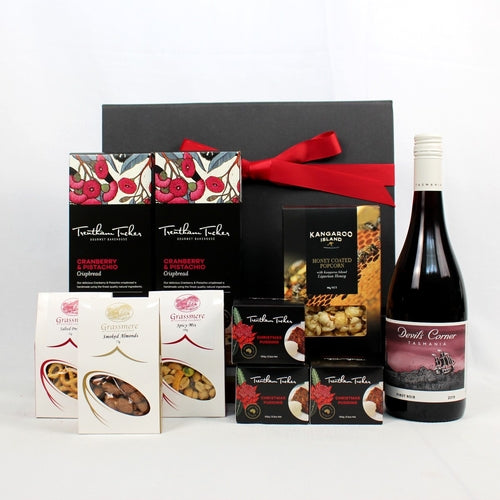 a black gift box with festive red ribbon standing behind a bottle of Australian red wine and a selection of sweet and savoury food like pistachio crispbread, pretzels, smoked almonds, spicy nut mix, honey glazed popcorn and three Christmas puddings