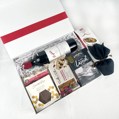 Open gift box with various items including a bottle of wine, a box of chocolates, and a red bow on a white background.