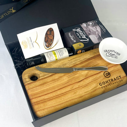 Wooden cutting board with knife, olive oil bottle, and snack in a gift box.
