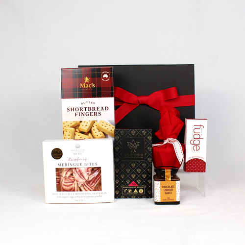 a black gift box with red ribbon behind a box of shortbread biscuits, meringue bites, chocolate bar, Christmas pudding in red fabric, chocolate pudding sauce and choc raspberry handmade fudge