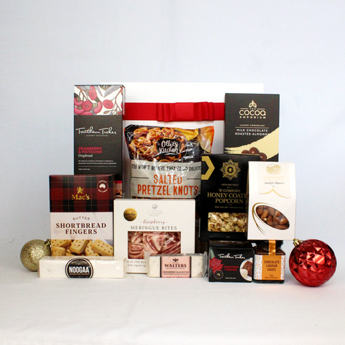 white gift box with red ribbon standing behind a range of sweet and savoury Christmas snacks including crispbread, shortbread, pretzels, nougat, raspberry meringue, honey coated popcorn, chocolate coated almonds, smoked almonds and a Christmas pudding with sauce