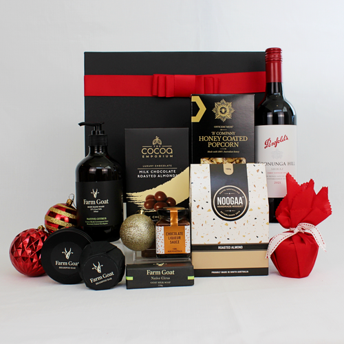 black gift box with red ribbon standing behind a bottle of red wine, christmas pudding and sauce, honey coated popcorn, nougat and chocolate coated almonds alongside ecosustainable hand wash, goats milk soap and shampoo bar