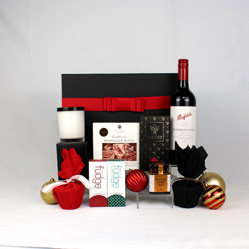 black gift box with red ribbon behind a bottle of Australian wine, luxury hand poured candle, raspberry meringue bites, chocolate bar, 2 small Christmas puddings, pudding sauce and 2 bars of handmade Australian fudge