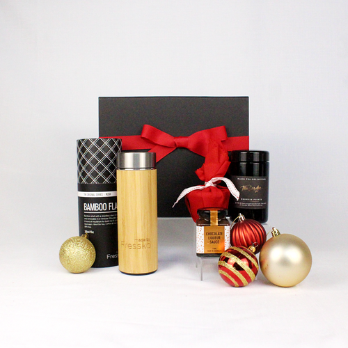 black gift box with festive red ribbon standing behind a reusable, sustainable bamboo drink flask accompanied by Christmas pudding in red fabric, pudding sauce and loose leaf tea surrounded by Christmas decorations