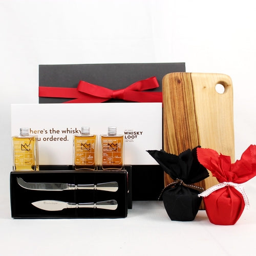 black gift box with festive red ribbon standing behind a whisky sample pack with 3 small whisky bottles, handmade timber cheese board with stainless steel cheese knife set and 2 Christmas puddings wrapped in black and red fabric