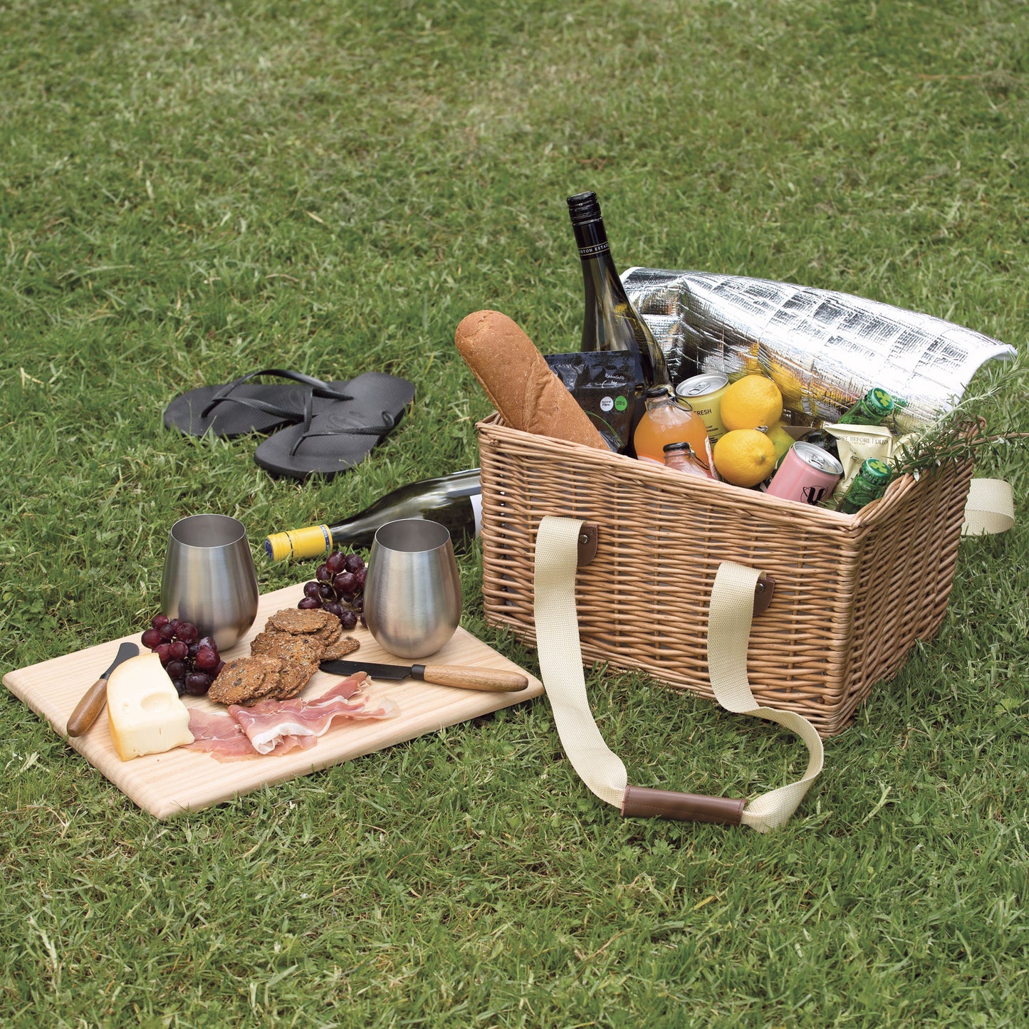 lifestyle image of wicker picnic basket opened and filled with a range of picnis fruits, drinks and bread next to a cheese board on the grass