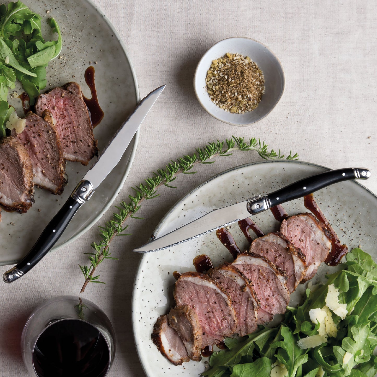 lifestyle photograph of black handled steak knives on plates with sliced meat, herbs and gravy