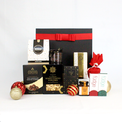 black gift box with red ribbon standing behind a range of sweet Christmas treats including Pudding and sauce, handmande chocolate, nougat, butter shortbread, chocolate coated almonds nad honey coated popcorn surrounded by Christmas bauble decorations