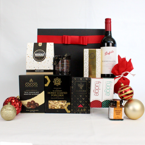 black gift box with red ribbon standing behind a bottle of Australian red wine and a range of sweet Christmas treats like Christmas pudding and sauce, fudge, handmade chocolate, chocolate coated almonds, nougat, shortbread cookies and surrounded by Christmas bauble decorations
