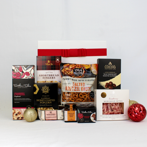 A white and red festive gift box standing behind a range of sweet and savoury festive snacks such as crispbread, pretzels, shortbread, chocolate coated almonds, honey coated popcorn, meringue, nougat, Christmas pudding with sauce