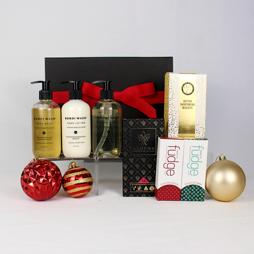 black gift box with red ribbon standing behind 3 bottle of handwash, body wash and hand lotion alongside some some delicious shortbread, fudge and chocolate
