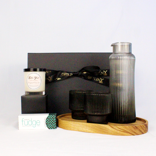 A luxury black magnetic gift box with ribbon filled iwth a handmade timber serving tray, mouth blown drinks carafe and glass pair, handpoured luxury candle jar and Australian made fudge.