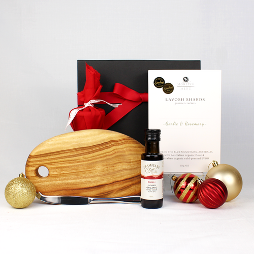 black gift box with red ribbon behind a small timber cheeseboard with single stainless steel knife, infused olive oil,  box of lavosh shards and a small Christmas pudding in red fabric surrounded by Christmas baubles