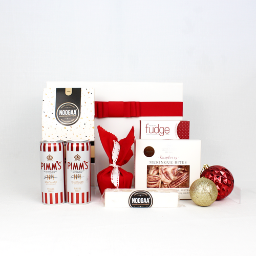 white gift box with red festive ribbon standing behind a box of nougat, chocolate fudge, meringue bites and a Christmas pudding in red fabric next to 2 cans of Pimm's premixed cocktails