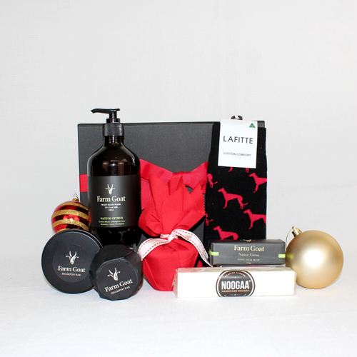 A black gift box with red ribbon standing behind a pair of corporate socks, a bottle of native citrus hand wash, shampoo bar and goats milk soap alongside a bar of nougat and a christmas pudding in red fabric