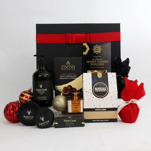 black gift box with red ribbon standing behind a range of Farm Goat bdy wash, shampoo bar and soap alongside 2 christmas puddings in red fabric with jar of pudding sauce, a  box of choocolate coated almonds, honey coated popcorn and nougat pieces