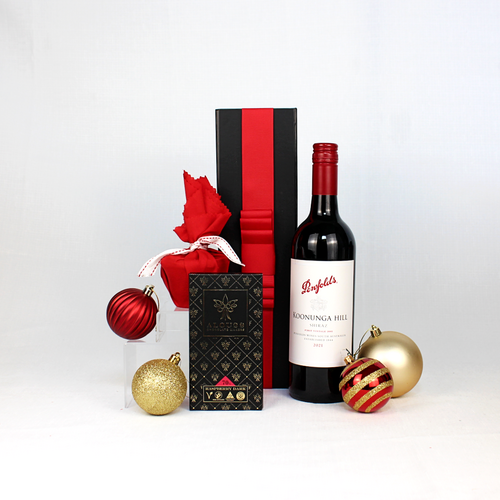 Black gift box with read ribbon behind a bottle of Australian red wine, Christmas pudding and handmade chocolate surrounded by Christmas baubles