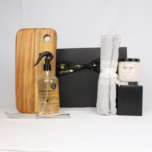 A black gift box with ribbon filled with beautiful home products such as a handmade timber cheese board with stainless  steel cheese knife, kitchen bench spray, luxury soy candle jar and a 100% cotton hand towel.