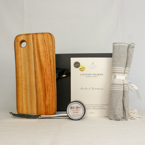 A handmade timber cheese board with stainless steel cheese knife, accompanied by lavosh shards, and cotton hand towel and a small luxury soy candle tin