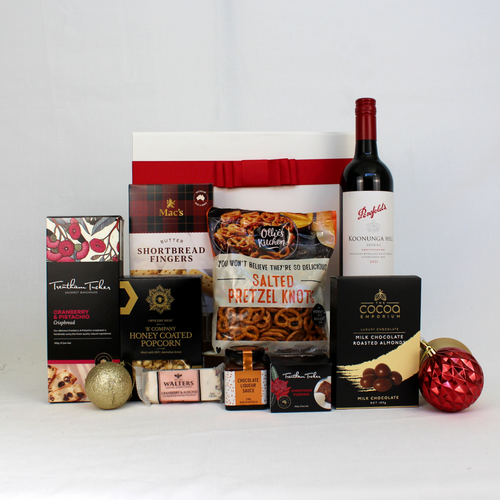 A white and red gift box behind a range of Christmas treats such as pudding and sauce, pretzels, crispbread, shortbread, chocolate coated almonds, nougat and a bottle of Australian red wine surrounded by Christmas baubles.