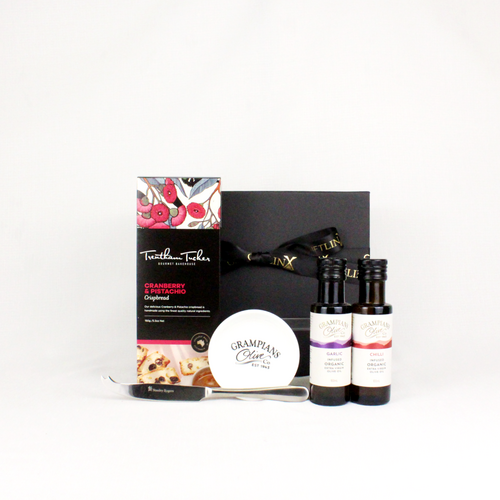 black gift box with ribbon behind a box of crispbread and 2 bottles of infused olive oil alongside a dipping bowl and cheese knife