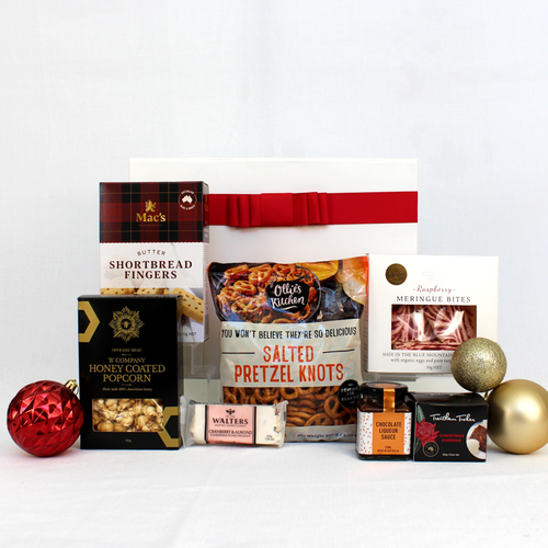 A white gift box with red ribbon behind a range of Christmas treats including pretzels, honey coated popcorn, shortbread, meringue, nougat and a Christmas pudding with sauce surrounded by decorative Christmas baubles