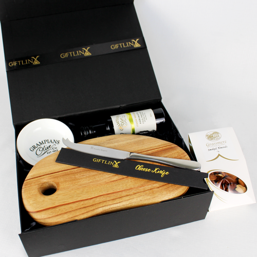 handmade timber serving boardwith cheese knife laying on top inside a black gift box alongside a bottle of olive oil and dipping bowl with a box of smoked almonds to the side.