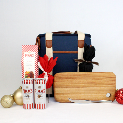 NAVY BLUE CANVAS COOLER BAGBEHIND A MEDIUM TIMBER CHEESE BOARD AND CHEESE KNIFE, 2 cans of premade Pimm's cocktails, a box of Pimm's chocolate truffles in a red and white striped box and 2 Christmas puddings wrapped in red fabric
