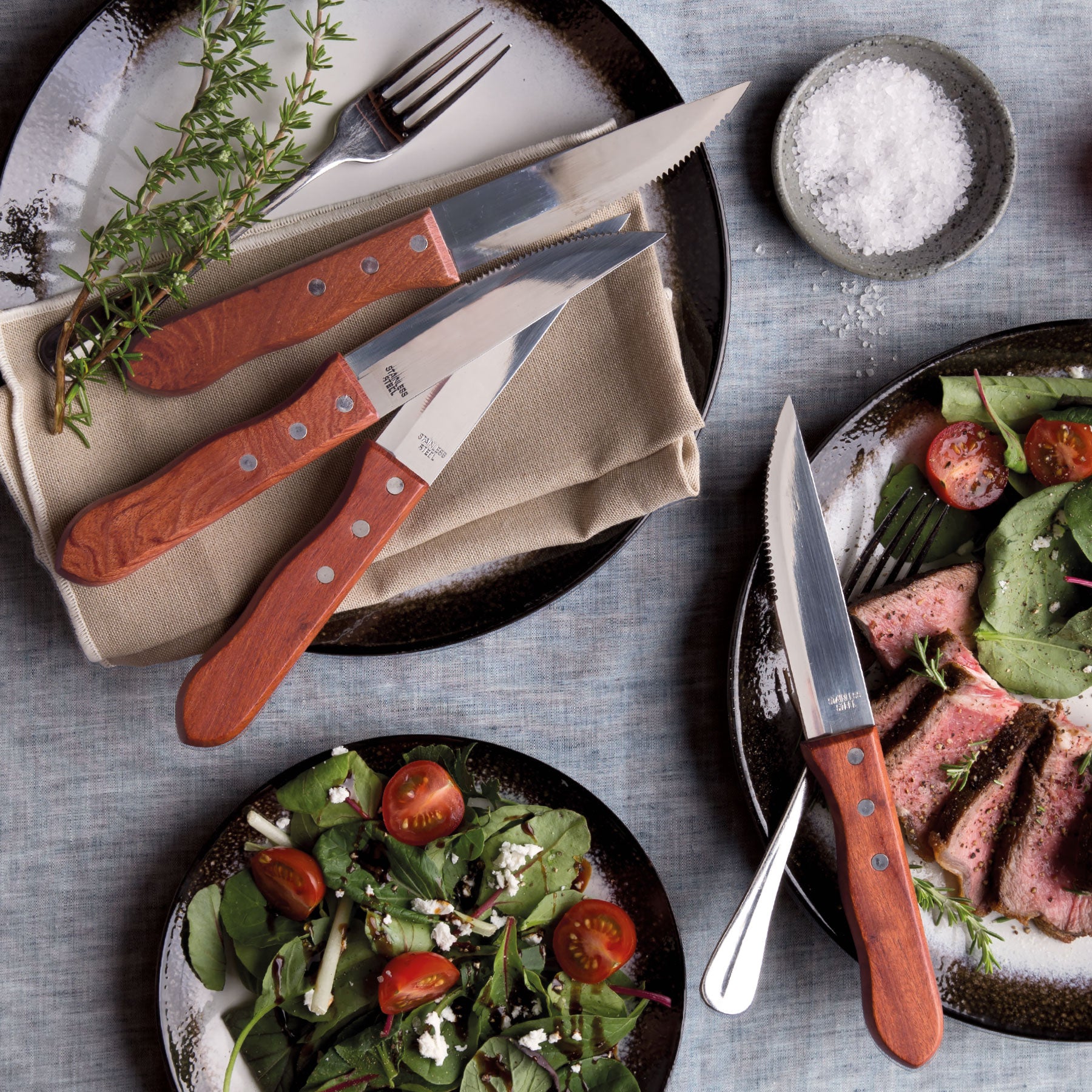 Lifestyle photo of 4 stainless steel blade steak knives with rosewood handles on top of black tableware with sliced meat and colourful salad