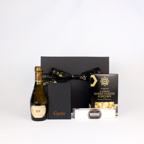 A piccolo of sparkling Australian wine next to black box of chocolate, honey coated popcorn and a bar of nougat all standing in front of a black gift box with ribbon