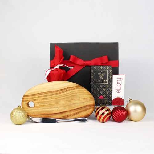 black gift box with red ribbon behind a handmade timber cheese board with stainless steel cheese knife plus Australian chocolate, fudge and Christmas pudding in red fabric with Christmas baubles surrounding the set up